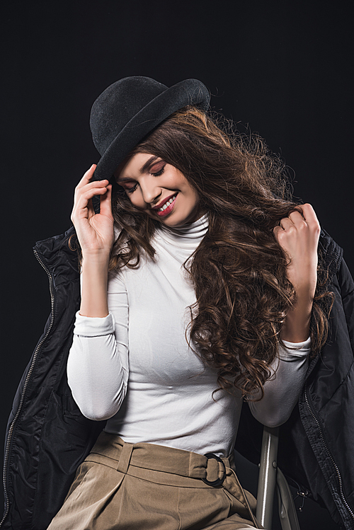 smiling young woman in stylish hat and winter jacket sitting on ladder isolated on black