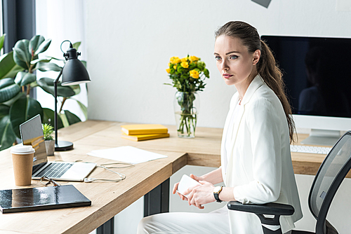 businesswoman in stylish suit with smartphone at workplace with laptop in office