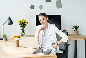 smiling beautiful businesswoman in white suit sitting at workplace with laptop and coffee to go in office
