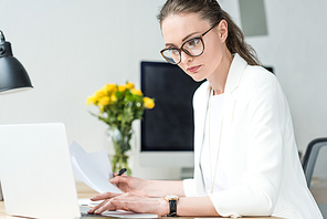 selective focus of businesswoman in eyeglasses with papers working on laptop at workplace in office