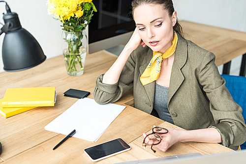 high angle view of beautiful stylish businesswoman holding eyeglasses and looking away while sitting at workplace