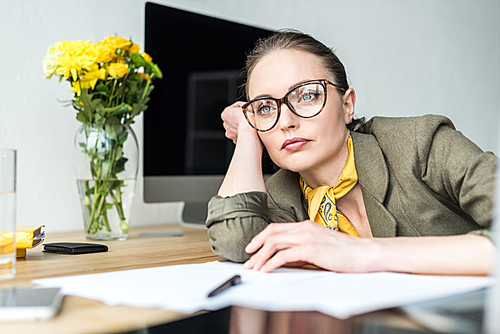 pensive businesswoman in eyeglasses leaning at table and looking away in office