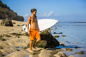 young surfer with surfing board standing on sandy beach near ocean