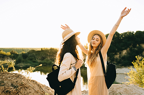 beautiful female friends in straw hats with backpacks having fun near lake with back light