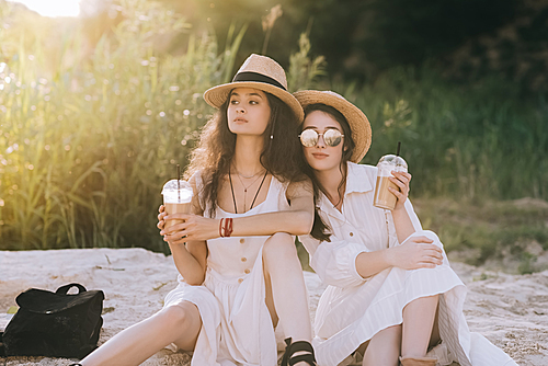 beautiful female friends in straw hats holding plastic cups with coffee latte and sitting on ground with sunlight
