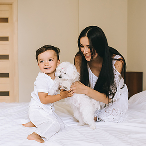 smiling young mother and child playing with bichon dog while sitting on bed at home
