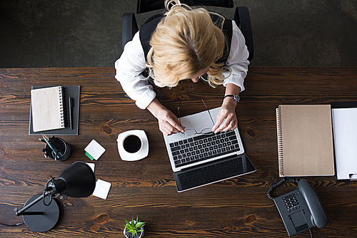 overhead view of businesswoman holding glasses above laptop in office