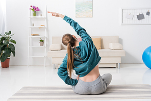 rear view of girl sitting in lotus position and stretching on yoga mat at home