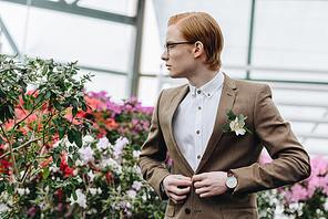handsome young groom in eyeglasses buttoning suit jacket and looking away in botanical garden