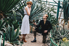 stylish young man standing on knee and making proposal to beautiful red-haired girl in botanical garden
