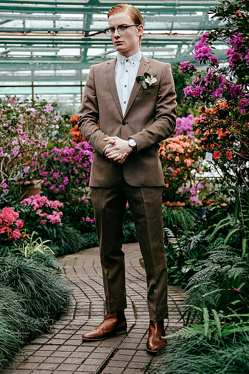 fashionable young man with red hair in suit and eyeglasses standing in glasshouse