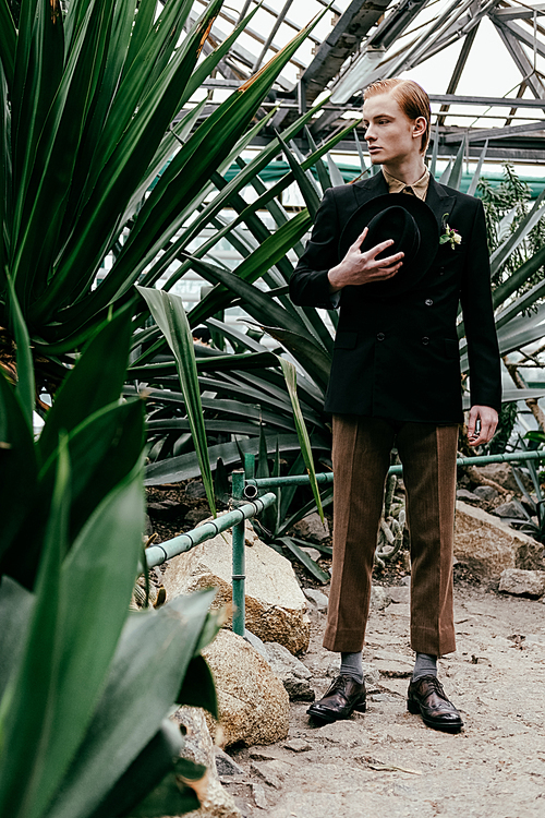 stylish young red hair man with hat in hand standing in greenhouse