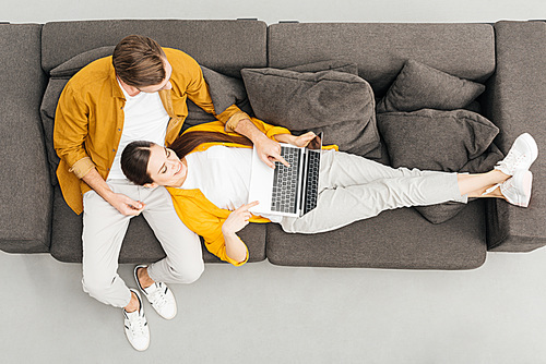 top view of woman lying on knees of boyfriend and working with laptop on cozy couch at home