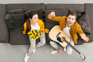 overhead view of man with guitar raising fists and shouting while his girlfriend holding bouquet on couch at home