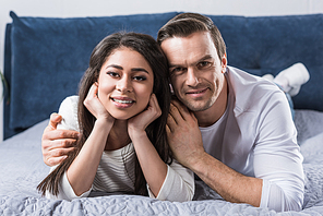 happy young multiethnic couple lying on bed and smiling at camera