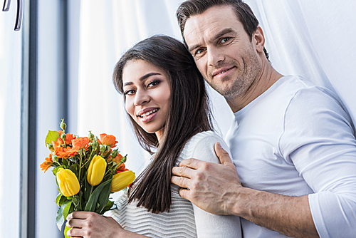 happy multiethnic couple embracing and smiling at camera while woman holding beautiful flower bouquet