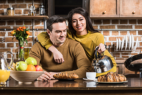 young african american woman holding kettle and smiling at camera while embracing boyfriend during breakfast at home