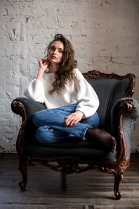 full length view of beautiful brunette woman in white sweater and jeans sitting on armchair and 