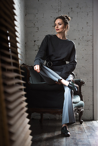 full length view of stylish young woman sitting on armchair and looking away indoors