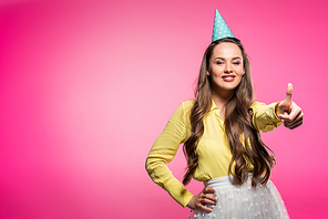 attractive woman with party hat showing thumb up isolated on pink