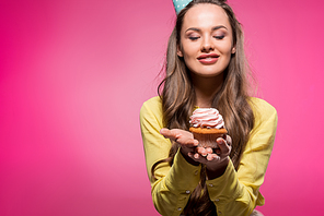 smiling attractive woman with party hat holding cupcake isolated on pink