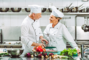 female and male chefs in uniform and hats looking at each other during cooking in restaurant kitchen