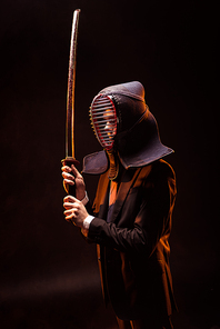 Kendo fighter in formal wear and helmet holding bamboo sword on dark