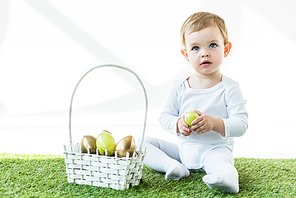 cute blonde baby holding yellow chicken egg while sitting near straw basket with Easter eggs  isolated on white