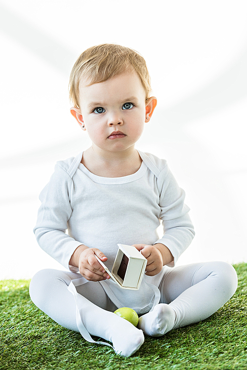 adorable kid sitting on green grass with white cardboard box and yellow chicken egg isolated on white