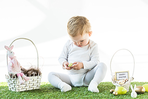 adorable child sitting near straw baskets with Easter eggs, decorative rabbits and happy Easter card isolated on white