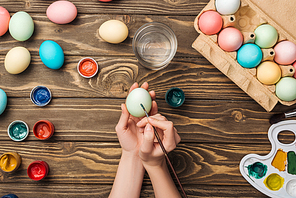 partial view of girl decorating easter eggs with paintbrush at wooden table with paints and color palette
