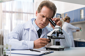 Concentrated scientist in white coat looking into the microscope in chemical laboratory