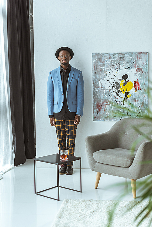 fashionable african american man in hat, checkered pants and blue jacket standing in modern office
