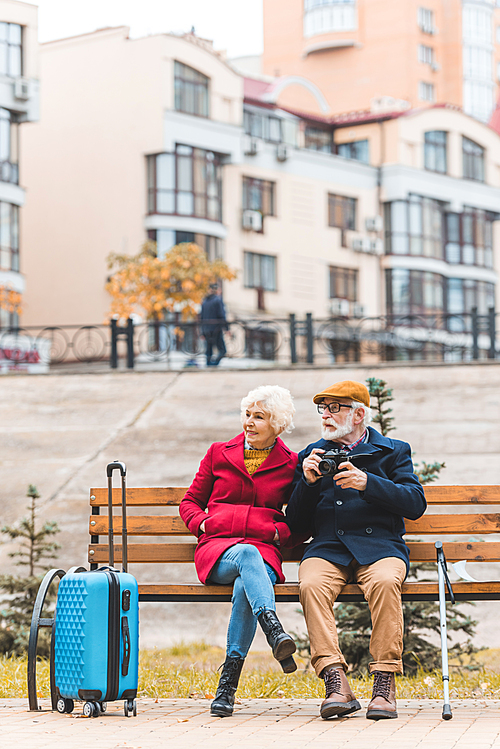 senior tourist couple with suitcase and photo camera sitting on bench in city