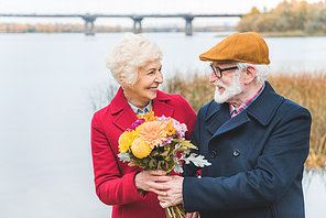 happy senior couple with bouquet of flowers standing at autumn lake