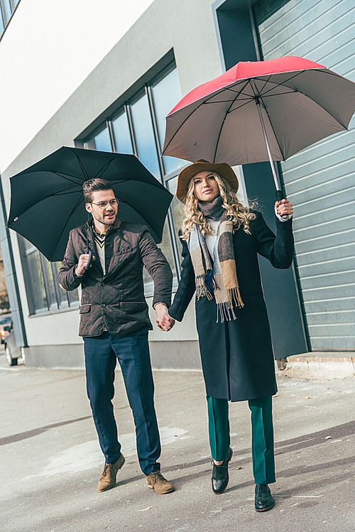 stylish young couple in autumn outfit holding umbrellas and walking together