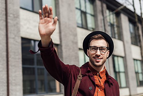 stylish young man in glasses and hat waving to someone