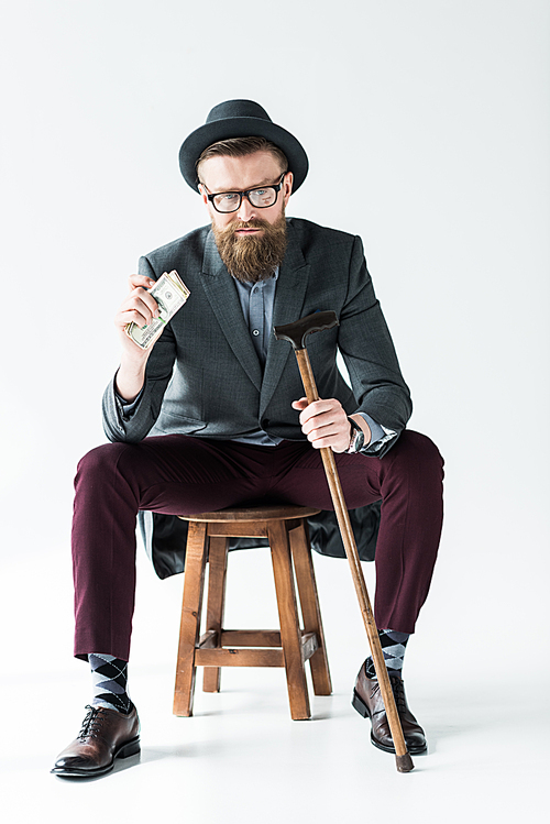 Stylish bearded businessman holding dollars and cane while sitting on stool