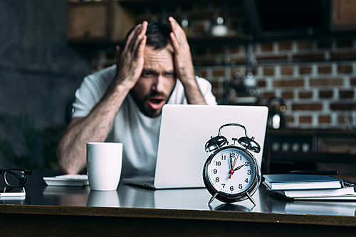 selective focus of stressed freelancer remote working at table with laptop and clock in kitchen at home