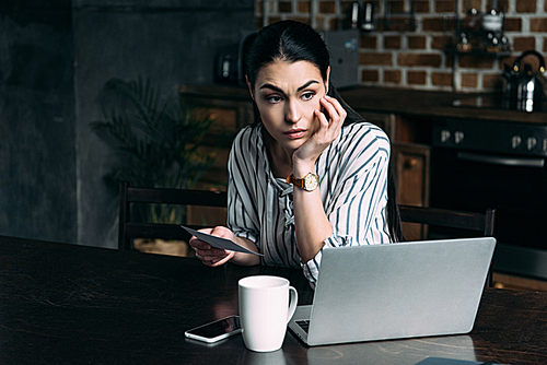 depressed young woman with smartphone and laptop sitting on kitchen and looking away