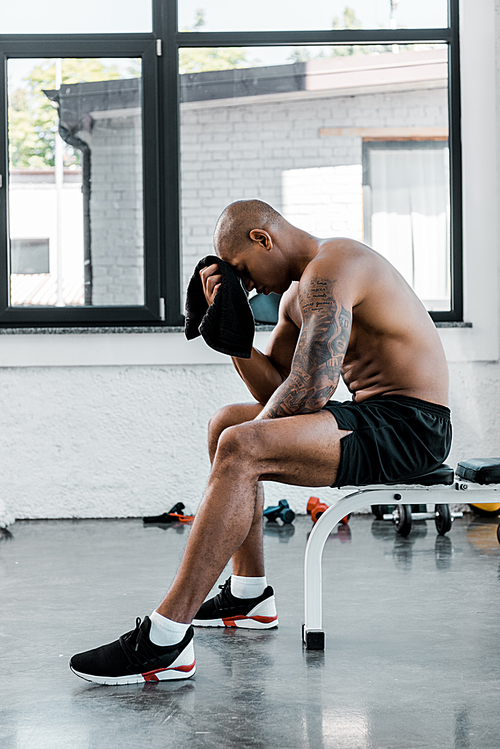 side view of muscular young sportsman wiping forehead with towel while sitting after workout in gym