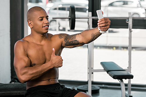 smiling muscular african american man showing thumb up and taking selfie with smartphone in gym