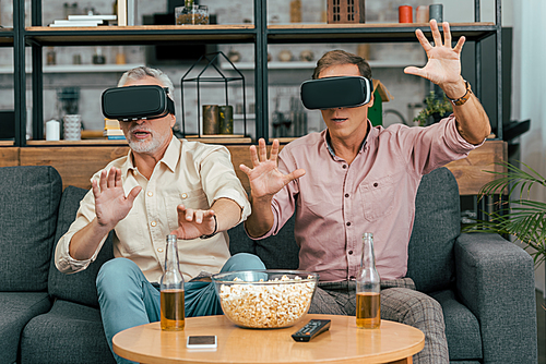 mature male friends using virtual reality headsets while drinking beer and eating popcorn at home