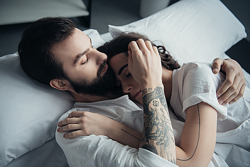 beautiful young tattooed couple embracing while lying in bed at home