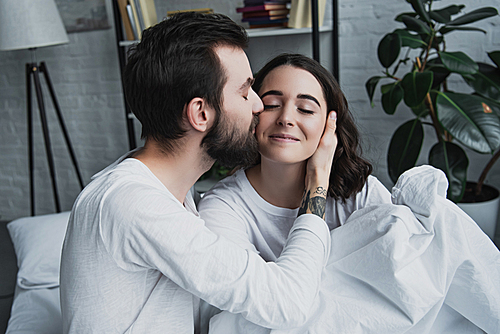 handsome bearded man kissing beautiful smiling woman at home in bed
