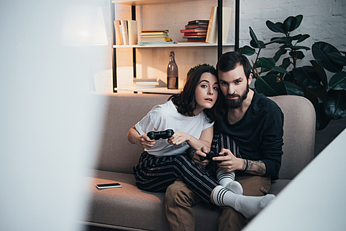 beautiful young couple sitting on couch with joysticks and playing video game in living room with copy space