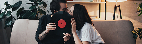 beautiful young couple covering faces with vinyl record and kissing while sitting on couch in living room