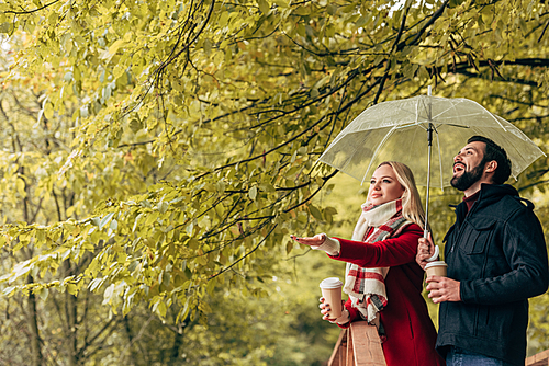 beautiful young couple holding umbrella and coffee to go in autumn park