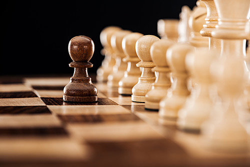 selective focus of wooden chessboard with beige chess figures and brown pawn in front isolated on black