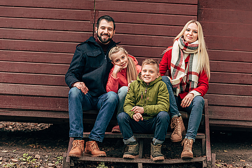 happy young family with two children sitting together on wooden stairs and smiling at camera